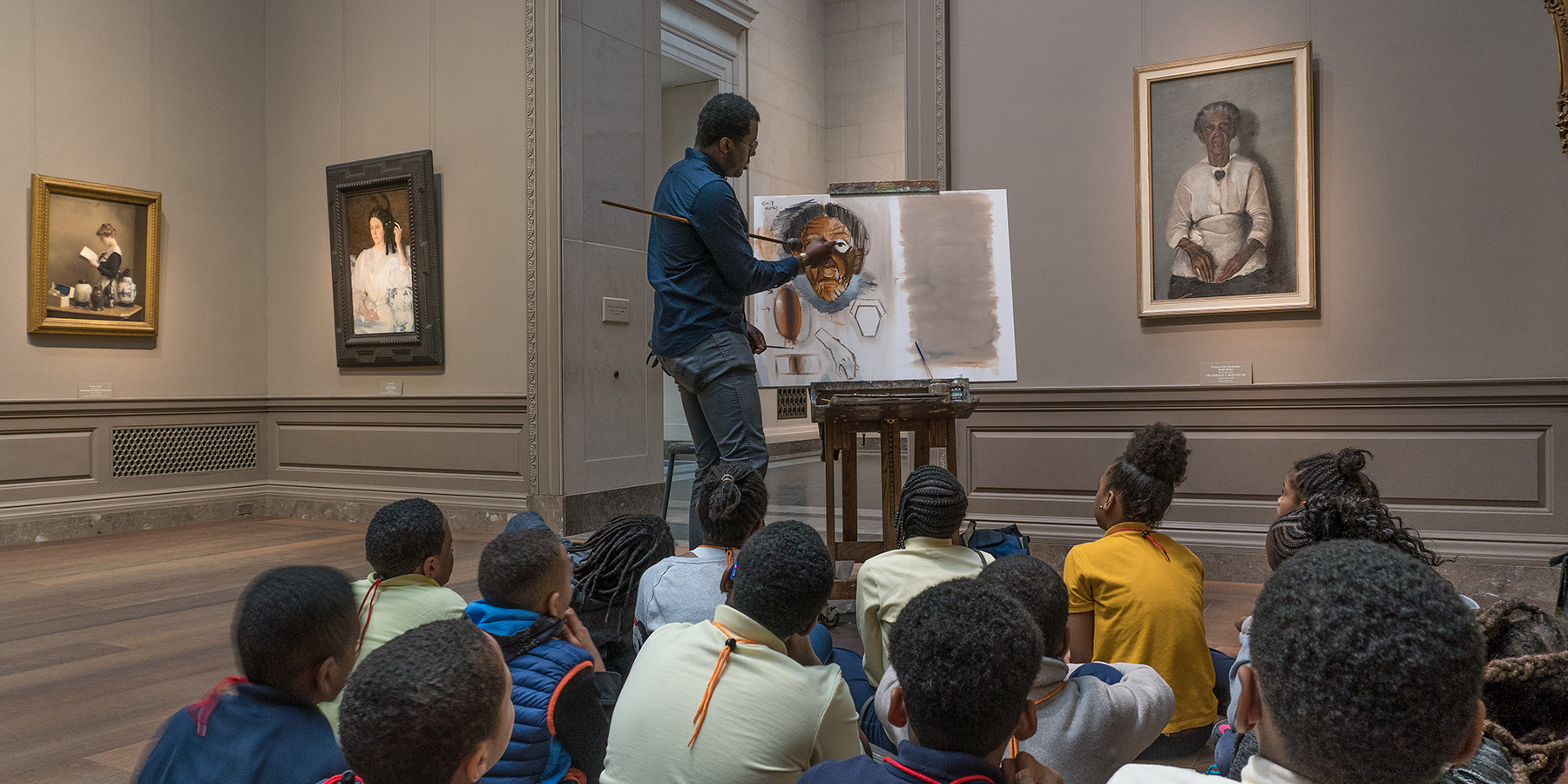 An art copyist is standing before a group of primary school aged children seated on the floor while he paints a copy of a portrait.