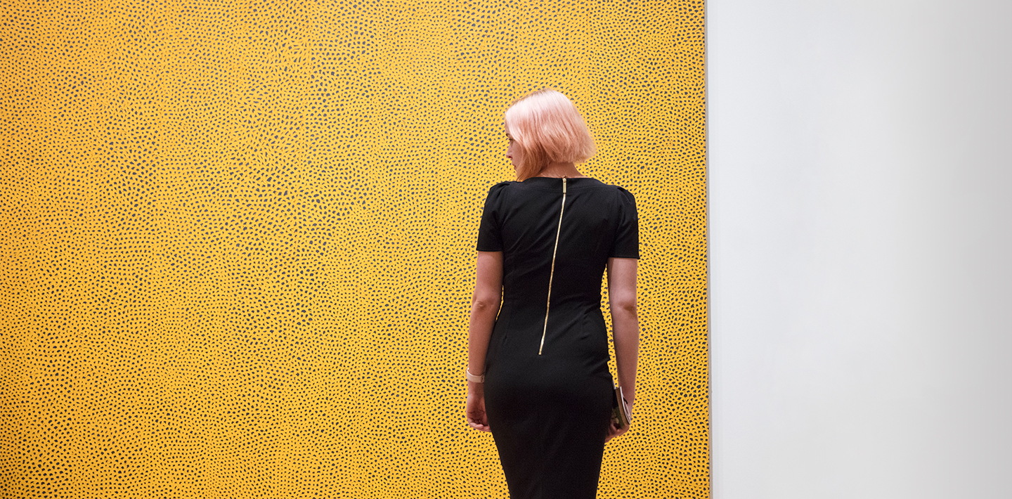 woman stands with her back to the viewer in front of a canvas that takes up the entire space of the image. The woman looks to her left. The canvas has a yellow and black abstract pattern on it. In contrast to the bright yellow of the work, the woman wears a black dress and has dull, pink hair.