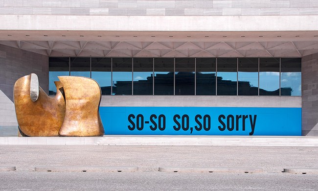 Front view of the East Building of National Gallery with alarge gold-toned sculpture by Henry Moore and Kate Rosen's SORRY painted on construction wall with the words so-so, so sorry.