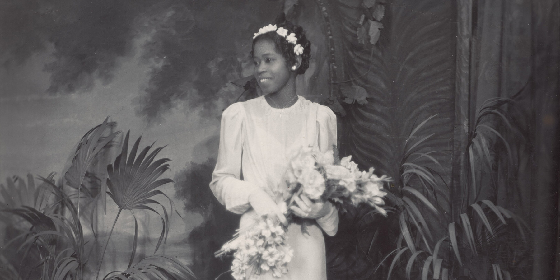 A woman in white wedding dress wearing a white flower headband holding a bouquet of flowers with palm plants behind her