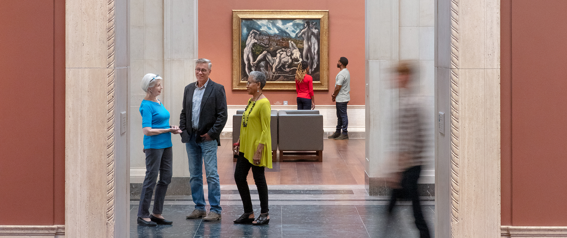Three people standing and talking inside the National Gallery of Art west building with a painting in the background and other visitors walking by