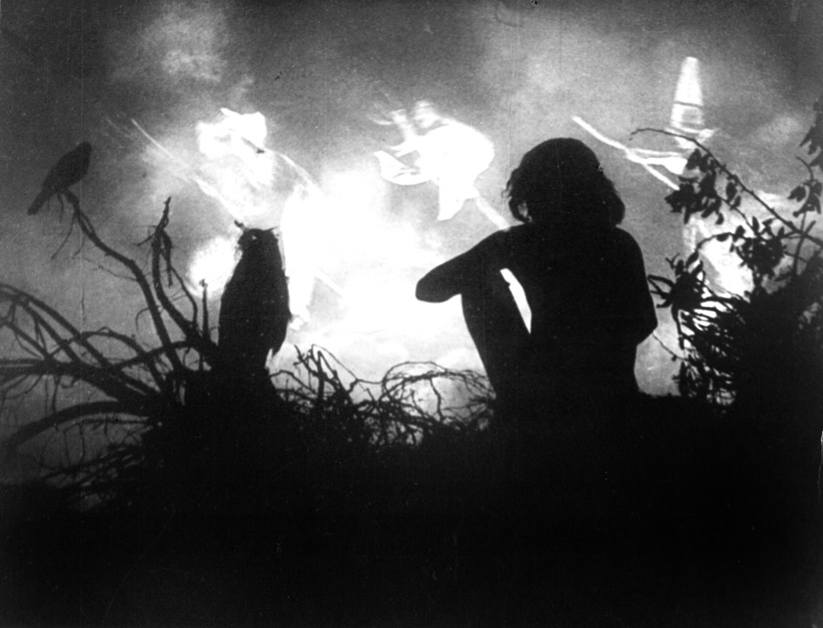 Black and white film still from Häxan (1922) showing a the siloutte of a seated person watching silouttes of three witches on broomsticks circling a fire.