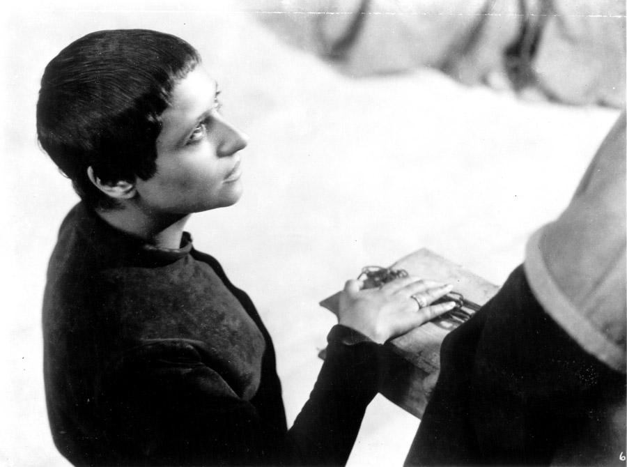 Black and white film still from The Passion of  Joan of Arc depicting a woman with short dark hair placing her hand on a book being held by a person wearing a robe that is partially off screen