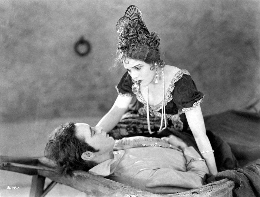 Black and white film still from Rosita (1923) showing a man laying on a cot and a woman looking at the man while seated on the far side of the cot