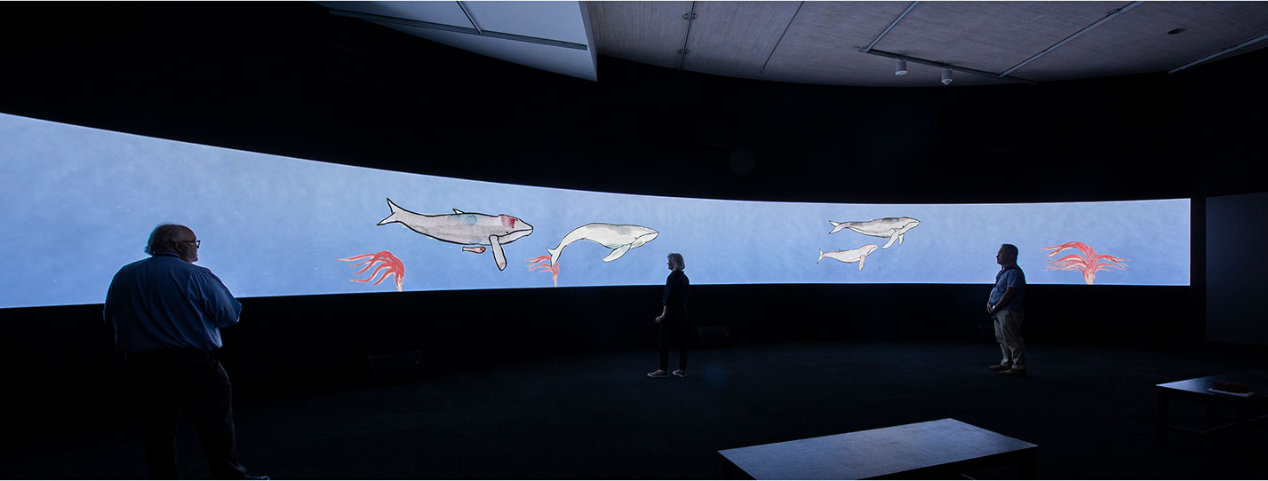Installation view of Avish Khebrehzadeh's video animation, "Seven Silent Songs"; projecyed in a darkened room on a wide, curvilinear screen. Animation depicts whales and octopi on a blue background.Three people are in the room, with their backs silhouetted against the screen.